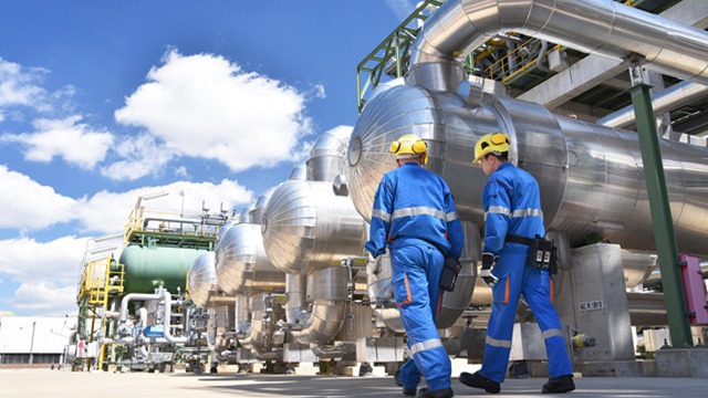 Two industrial workers in blue uniforms and yellow helmets inspect large metal pipes and machinery outdoors.
